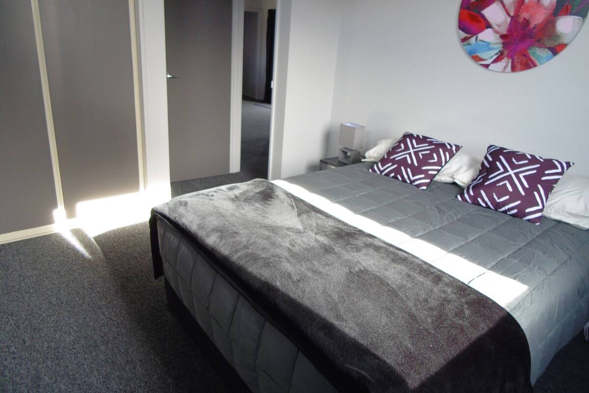 Ein modernes Schlafzimmer mit einem großen Bett, einem grauen, bezogenen Kopfteil und einem farbenfrohen, abstrakten Gemälde an der Wand.