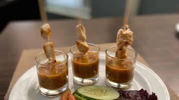Un piatto con tre piccoli bicchieri contenenti un liquido bruno, probabilmente una zuppa o salsa, accompagnati da una fetta di cetriolo, una fetta di pomodoro e verdure a foglia.