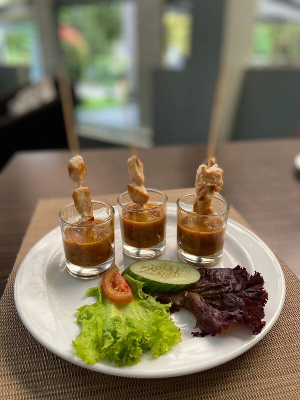 Un plato con tres vasos pequeños de un líquido marrón, posiblemente sopa o salsa, acompañado de una rodaja de pepino, una rodaja de tomate y algunas hojas verdes.