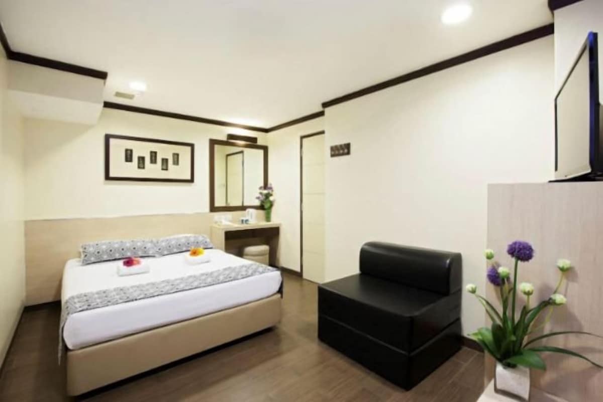 A hotel room with a bed, a desk, a black leather sofa, a mirror, and a potted plant.