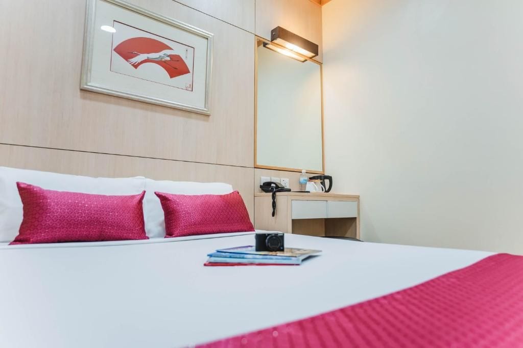 A hotel room with a bed, pillows, a bedside table with a camera, a mirror, and a framed picture on the wall.