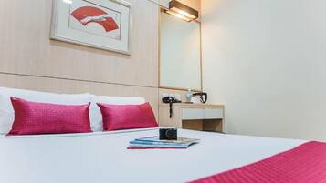 A hotel room with a bed, pillows, a bedside table with a camera, a mirror, and a framed picture on the wall.