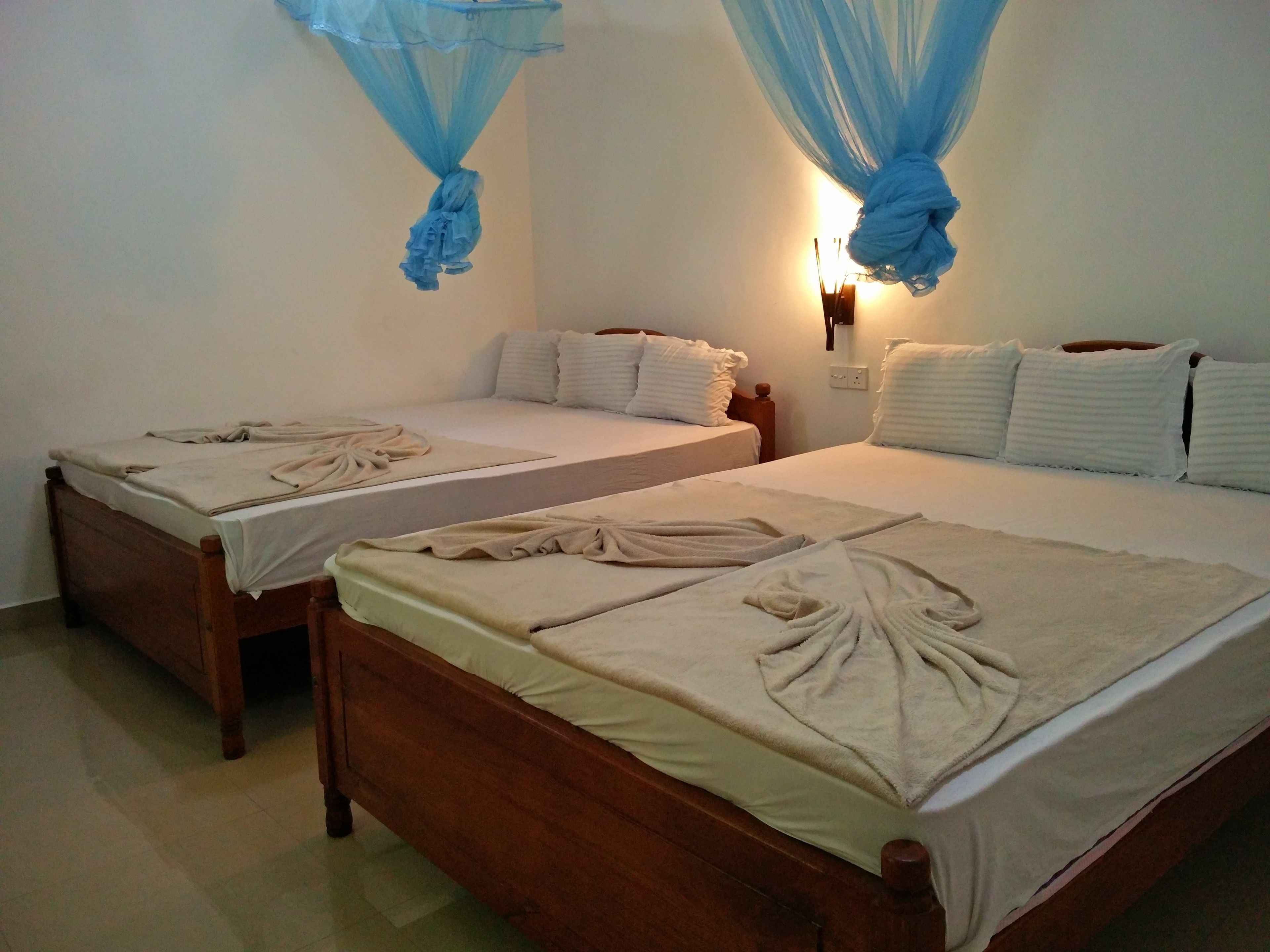 A room with two wooden beds, each with white bedding and a decorative towel. Blue mosquito nets hang above the beds.