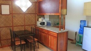 Een kleine keuken met houten keukenkastjes, een magnetron en een spoelbak. Er staat een eettafel met stoelen en een waterdispenser in de hoek.