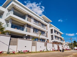 Un immeuble moderne à plusieurs étages, doté de balcons, d’une entrée sécurisée et d’un jardin bien entretenu agrémenté de fleurs rouges.