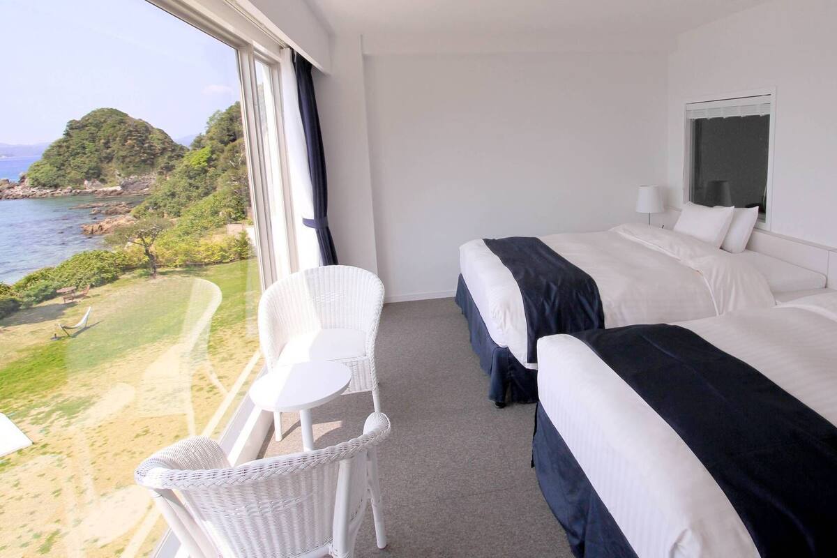 Ein Hotelzimmer mit zwei Betten, einem kleinen Balkonstuhl und Blick auf das Meer und Grünflächen.
