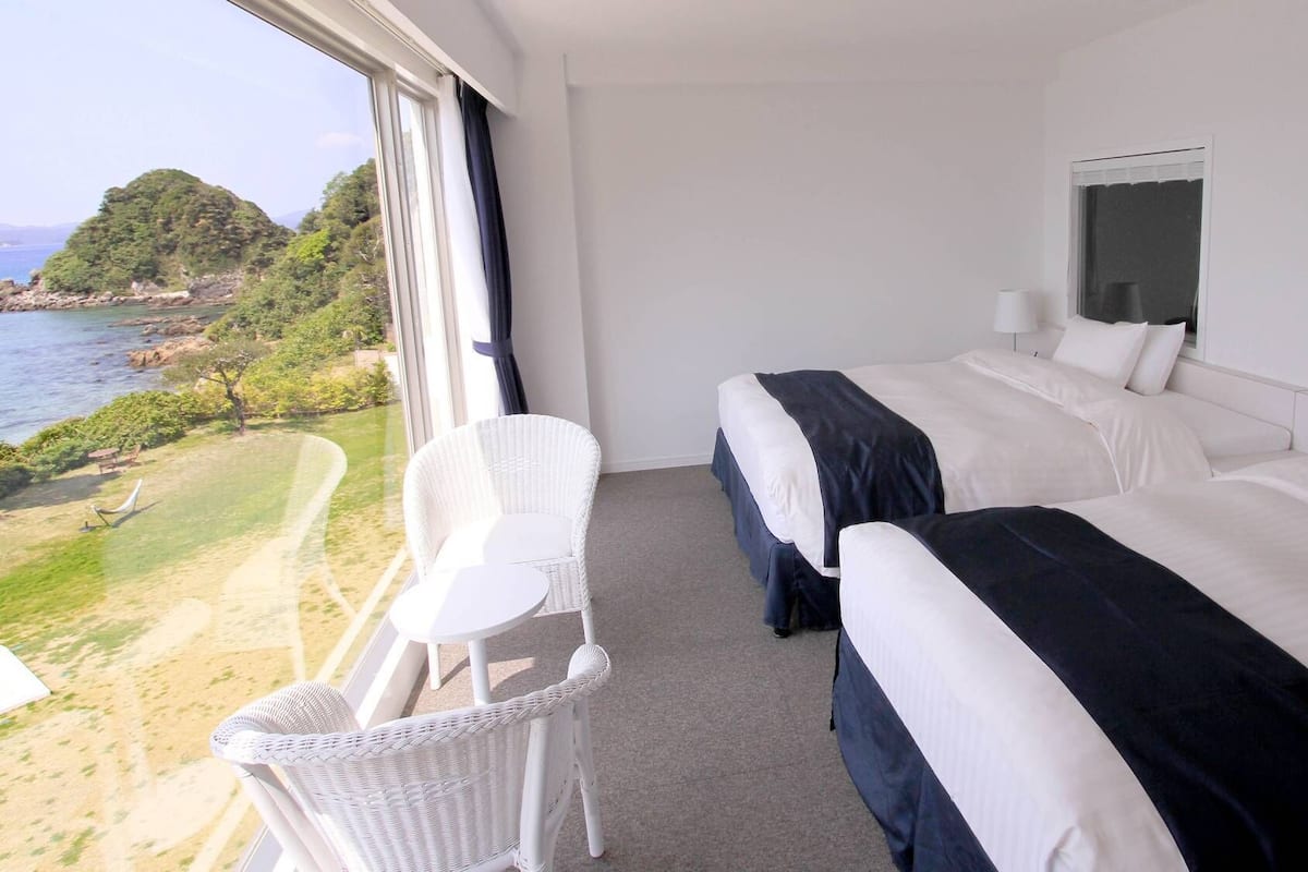 Une chambre d’hôtel avec deux lits, une petite chaise sur le balcon et une vue sur la mer et la verdure.