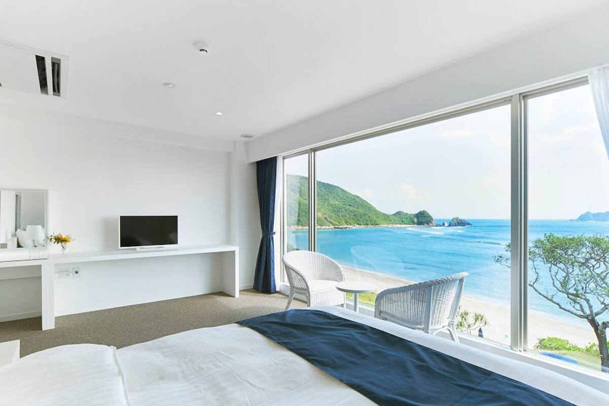 Ein modernes Hotelzimmer mit einem großen Bett, einem Fernseher auf einer weißen Kommode, einem Balkon mit einem Sessel und Blick auf das Meer und die Hügel.
