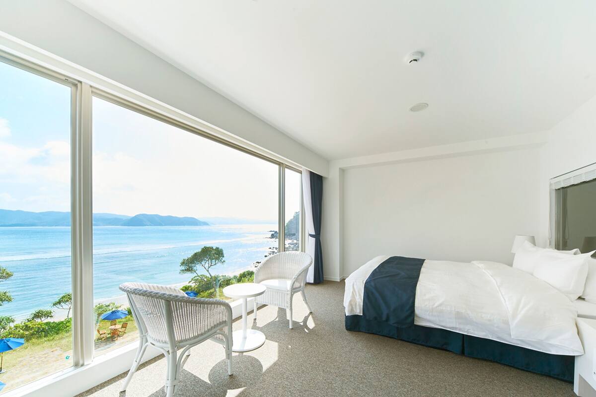 Ein modernes Hotelzimmer mit großem Fenster und Meerblick, einem Bett mit weißer Bettwäsche, einem kleinen runden Tisch und einem Korbstuhl.