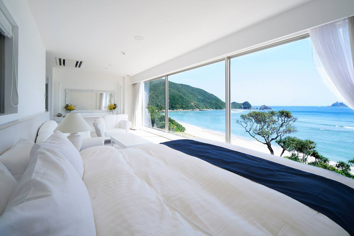 Ein modernes Schlafzimmer mit einem großen Bett, einem Nachttisch, einer Lampe und Blick auf das Meer und die Hügel.