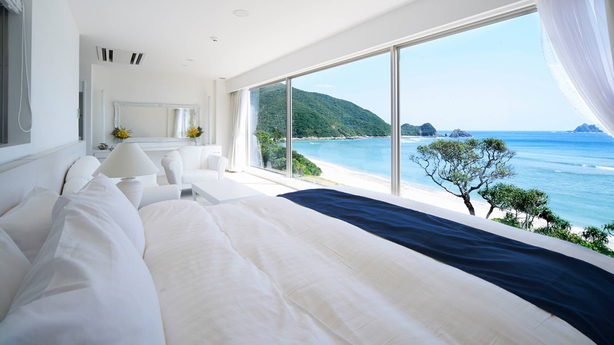 A bedroom with a large bed, a nightstand, a lamp, and a view of the ocean and a mountain.