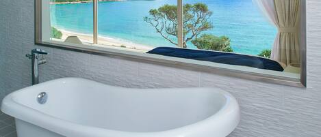 A bathroom with a bathtub, a window with a view of a beach and the sea, and a towel rack.