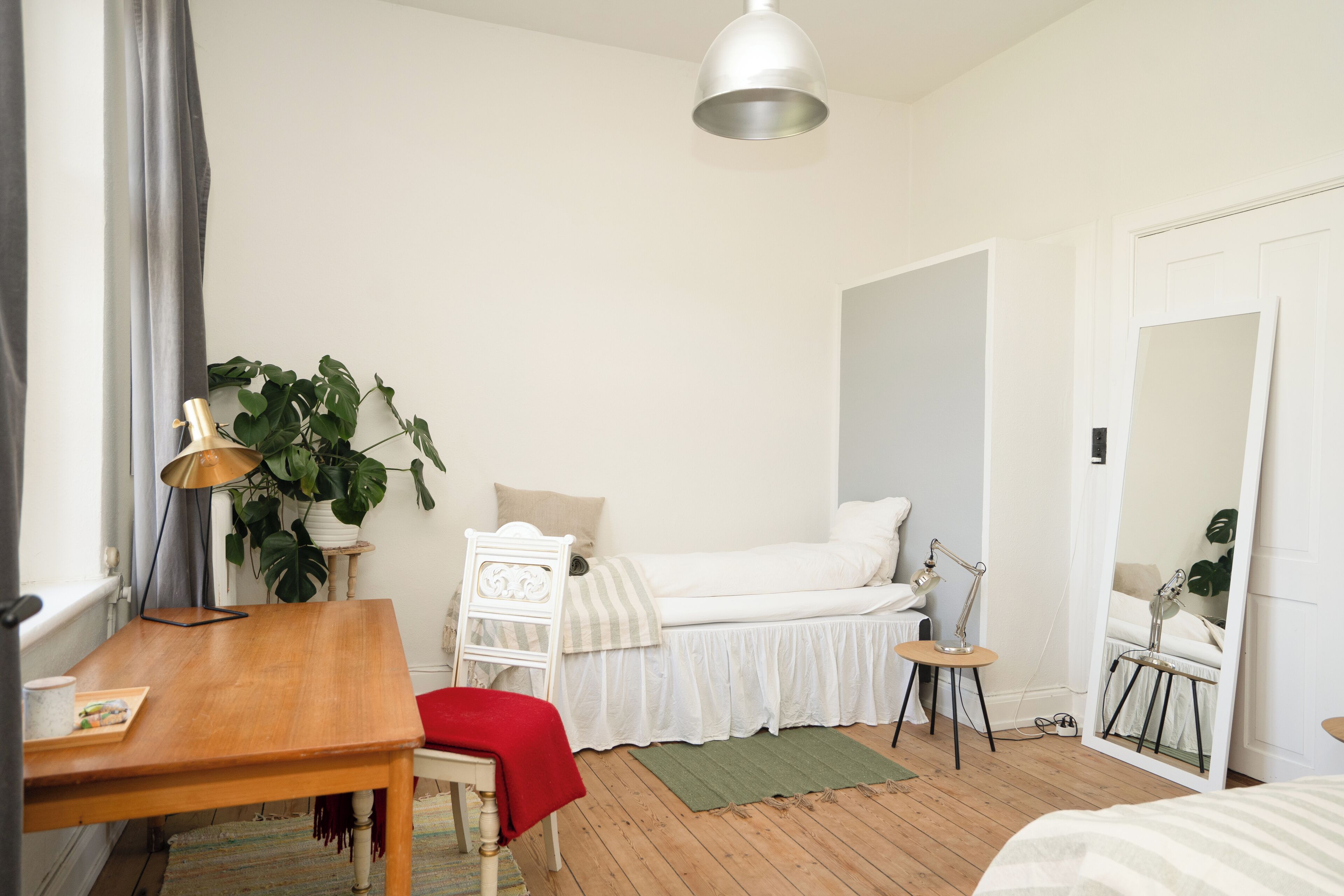 Ein Schlafzimmer mit einem hölzernen Bett, einem Nachttisch, einem Schreibtisch, einem Stuhl, einer Lampe, einem Spiegel und einer Topfpflanze.
