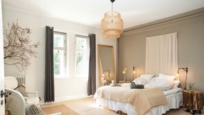 A bedroom with a large bed, a chair, a bedside table, a mirror, and a window with curtains.