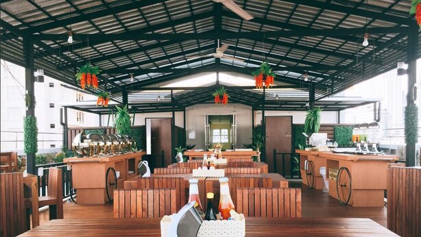 A spacious outdoor seating area with wooden benches and tables, surrounded by greenery and a metal roof structure.