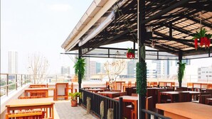 An outdoor seating area with wooden tables and chairs, potted plants, and a view of the cityscape.