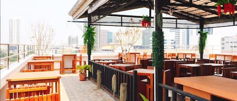 An outdoor seating area with wooden tables and chairs, potted plants, and a view of the cityscape.