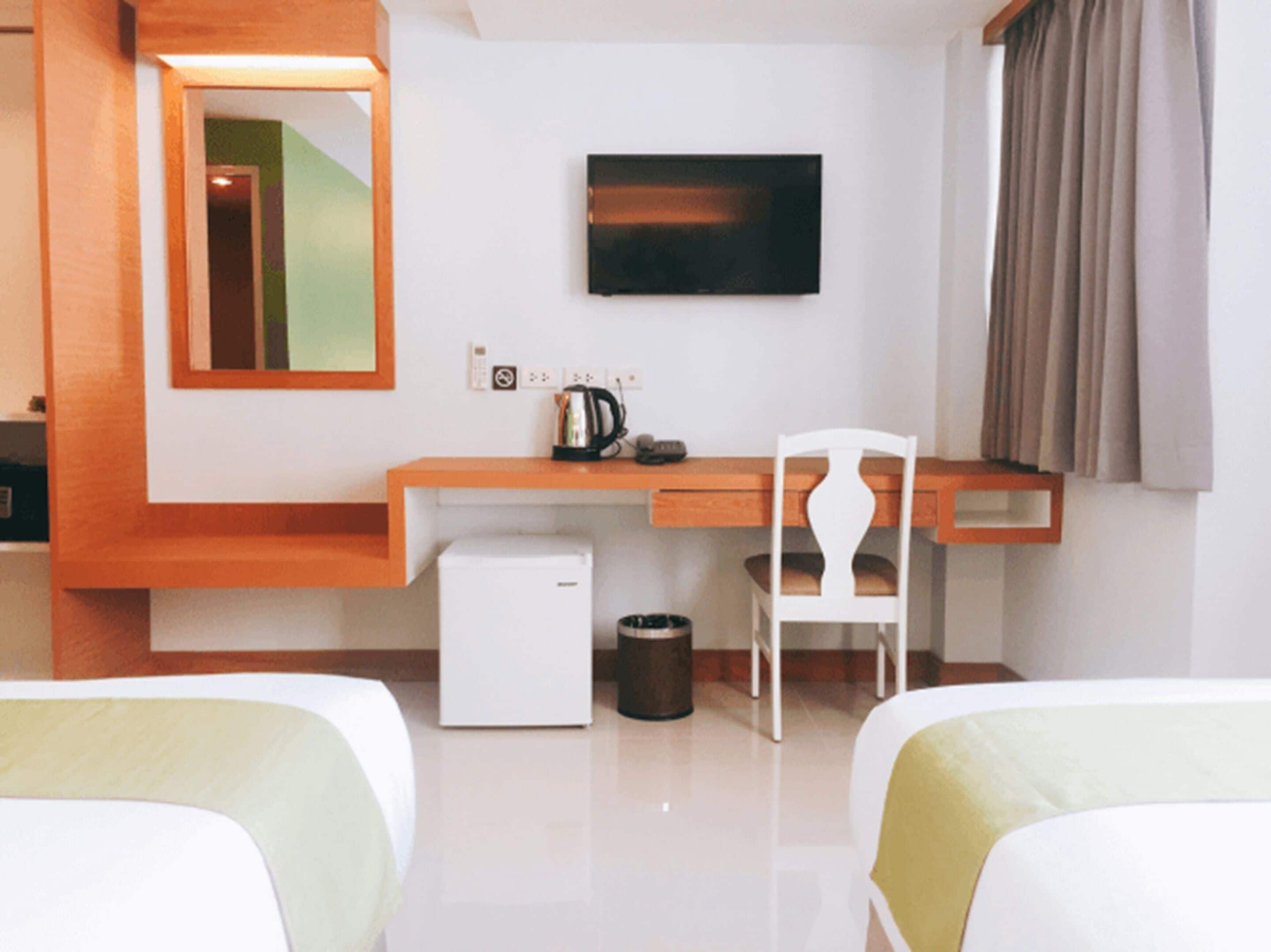 Habitación de hotel con un escritorio de madera, un refrigerador pequeño, una silla blanca, un televisor montado en la pared y dos camas con ropa blanca.