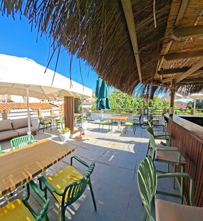 Eine Terrasse im Freien mit Tischen und Stühlen, einem Reetdach und Blick auf die Umgebung.