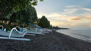 A row of boats on a pebbled beach with trees overhead and a calm sea in the background. - Akasa Villa Tulamben (Tulamben)