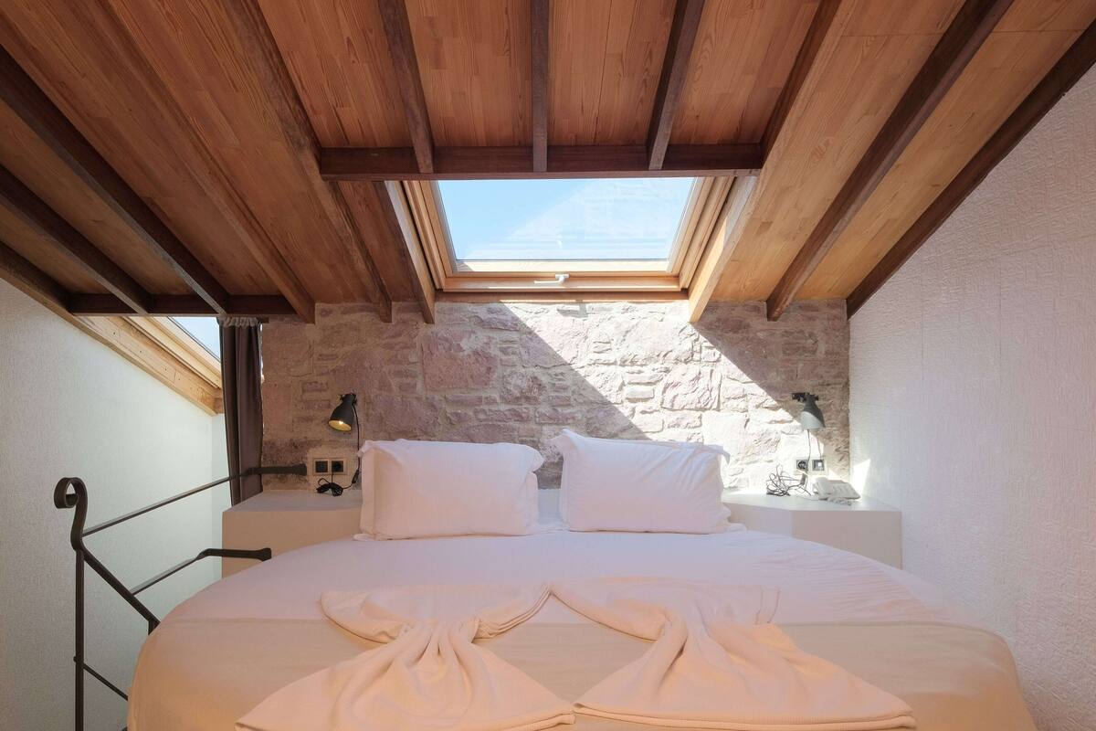 A bedroom with a skylight, stone walls, a bed with white linens, and a bedside table with a lamp.