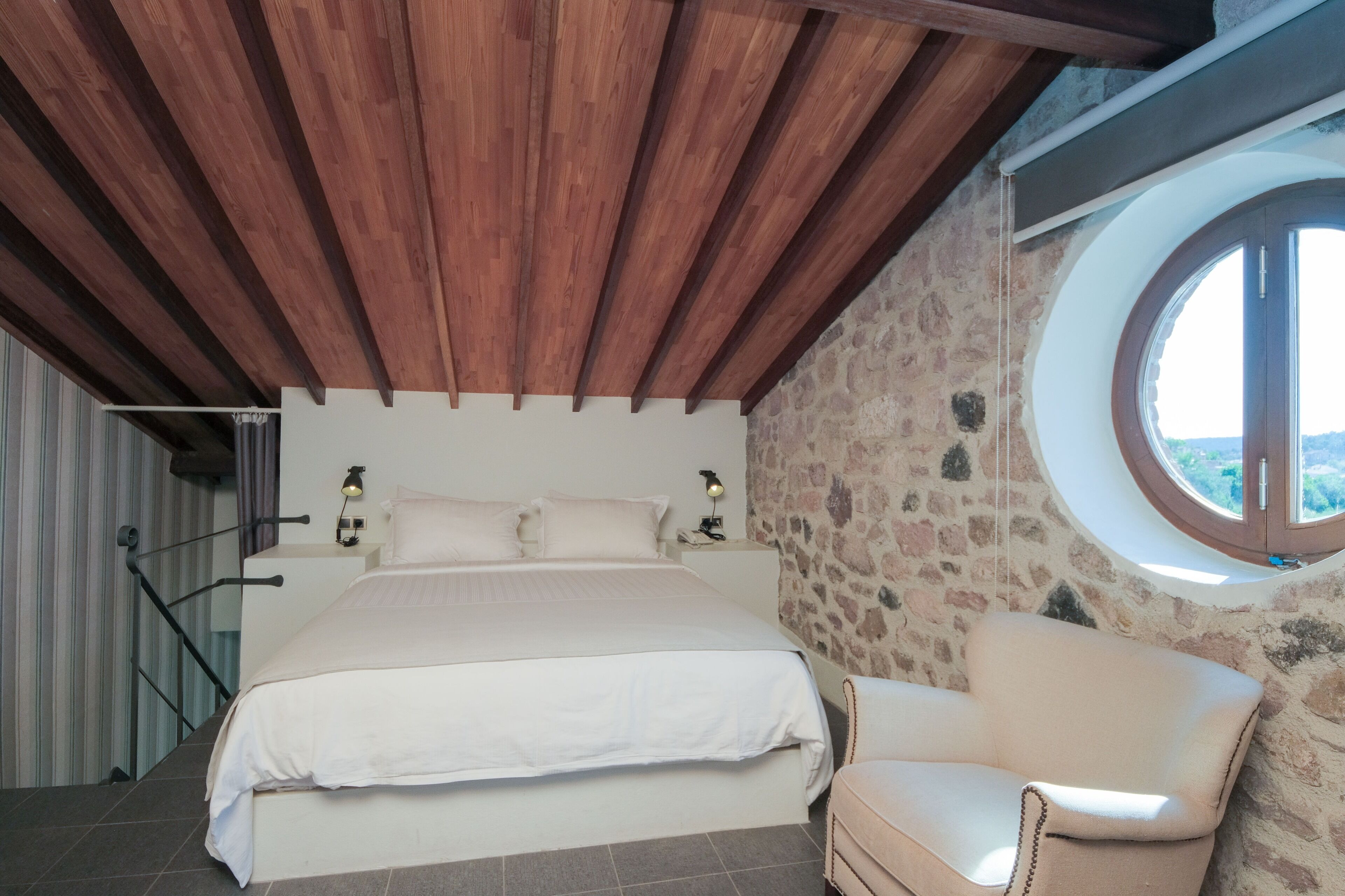 A bedroom with a large bed, a stone wall, a round window, and a wooden ceiling.
