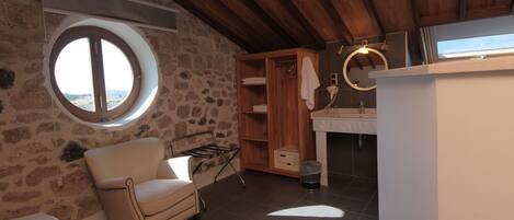 A rustic bathroom with a stone wall, wooden beams, a round window, a wooden cabinet, a sink, and a mirror.