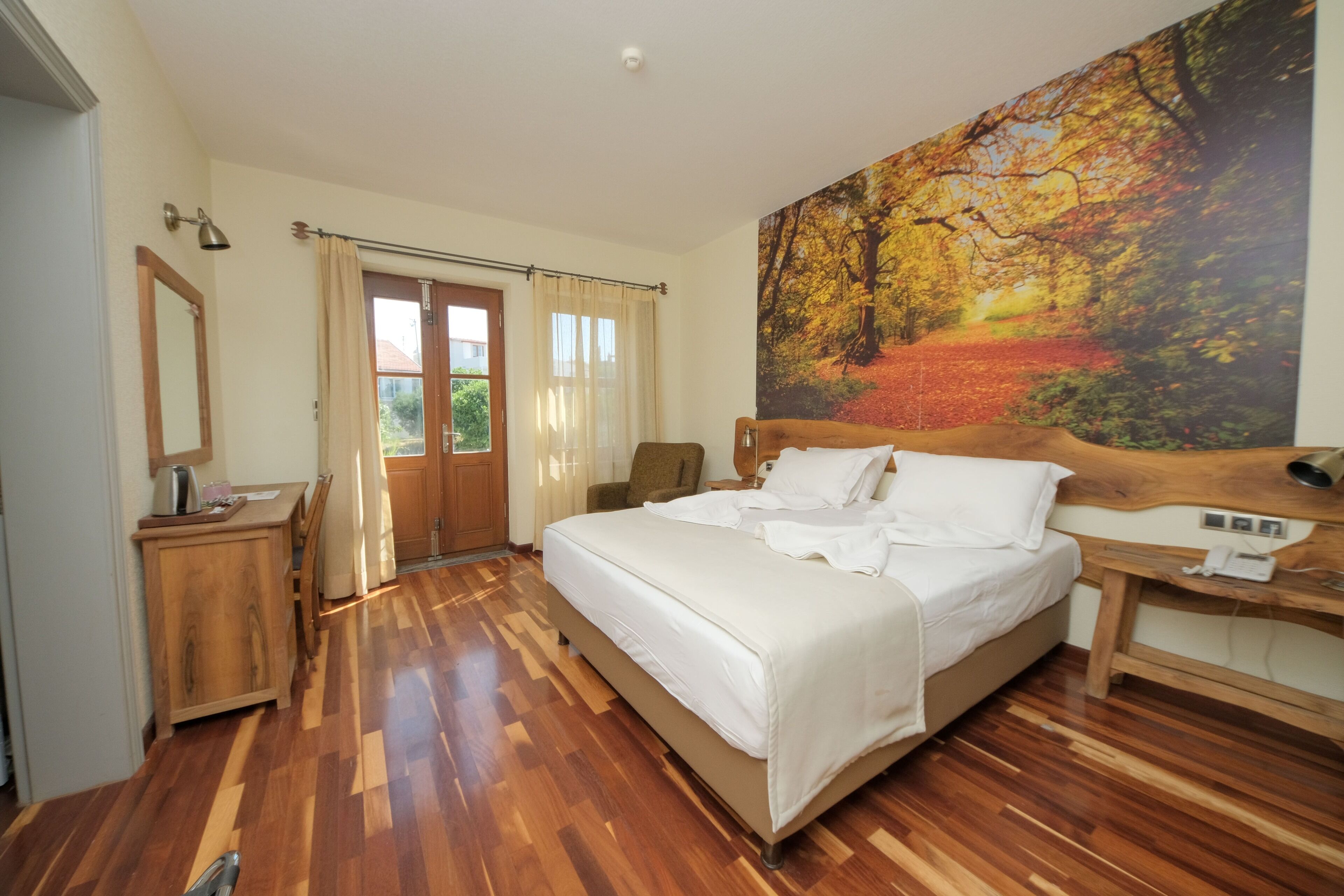 A bedroom with a large bed, wooden flooring, a painting of a forest, a wooden desk, and a chair.