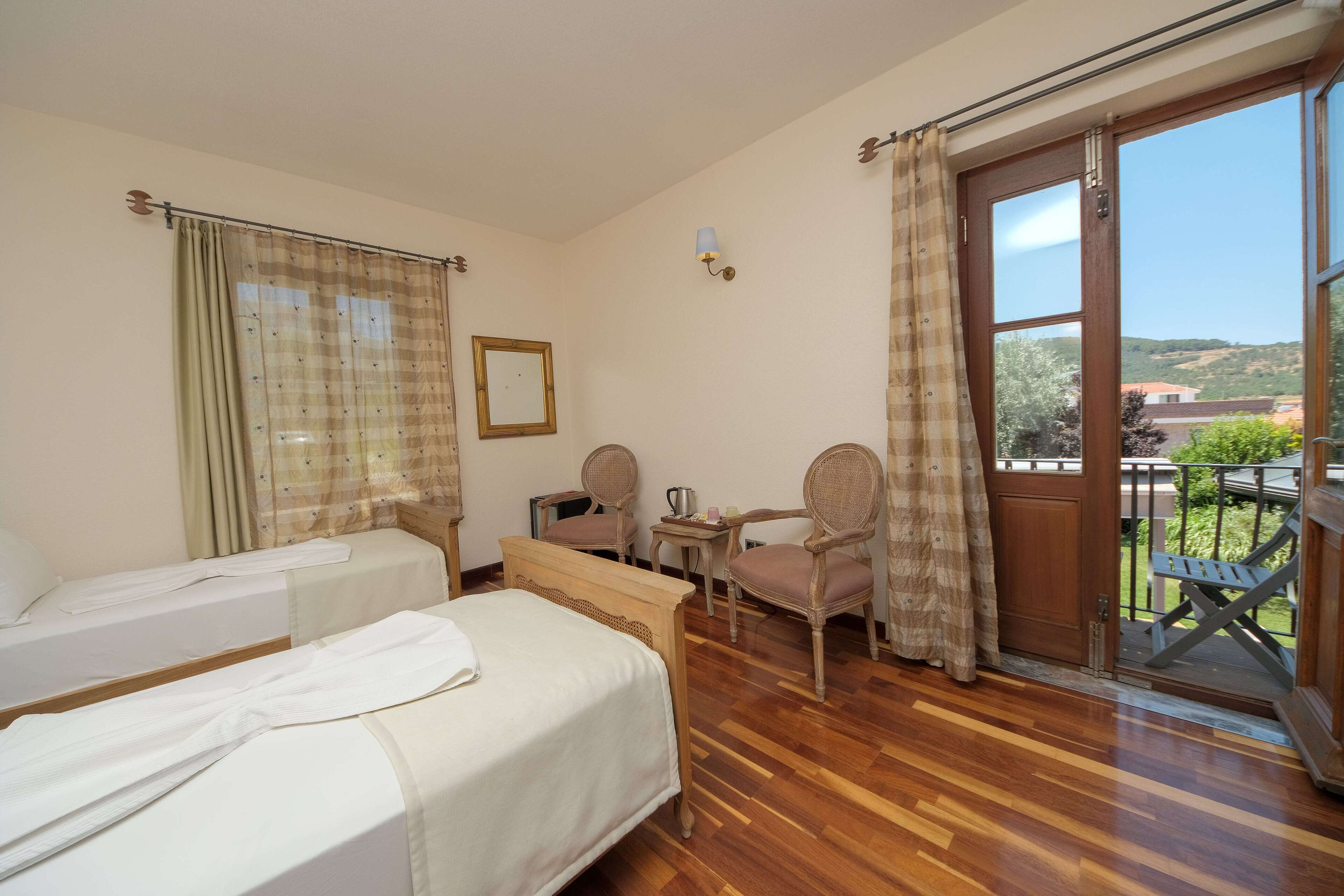 Habitación de hotel con dos camas, un banco de madera, una mesita, dos sillas y un balcón con vistas al campo.