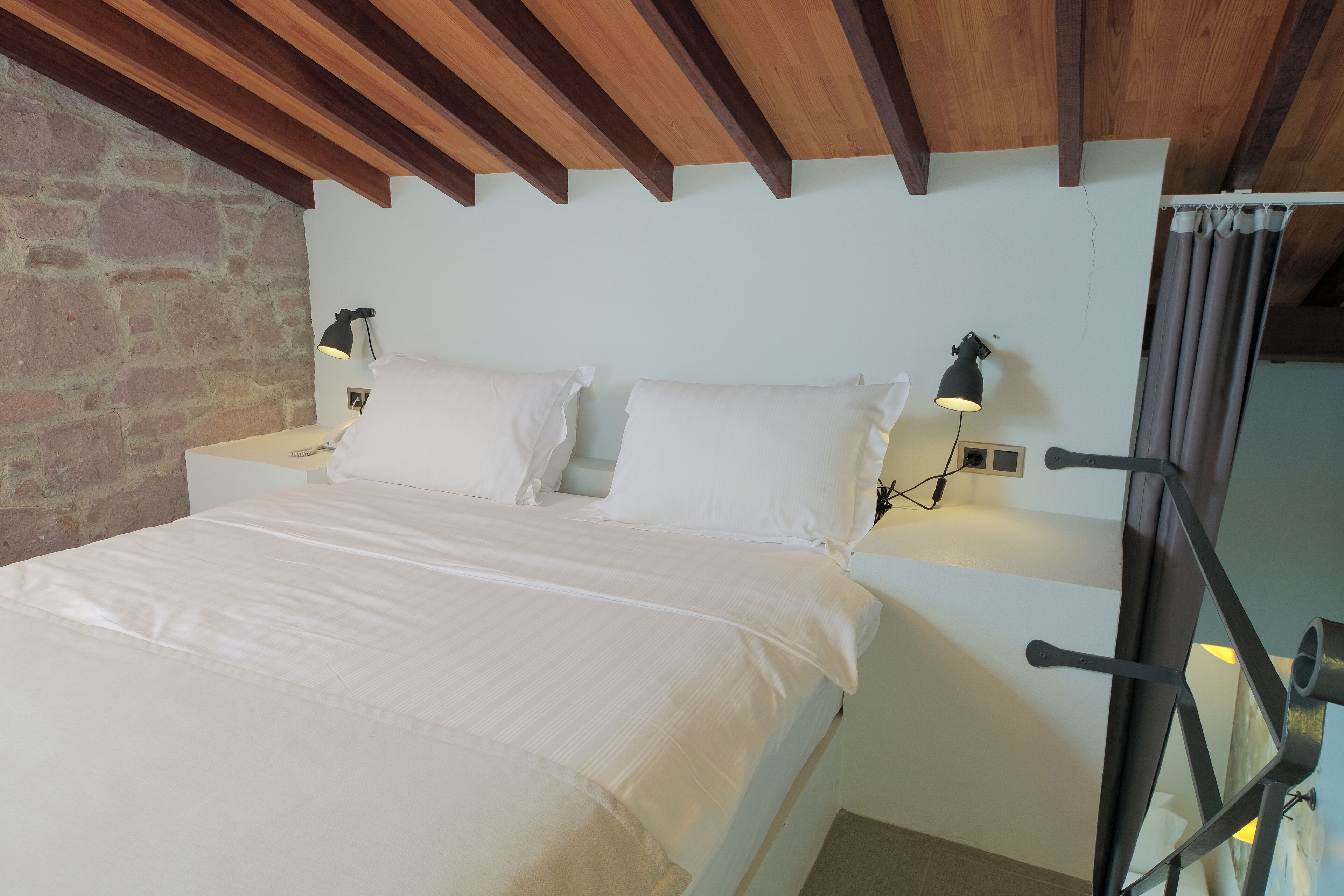 A neatly made bed with white linens in a room with exposed wooden beams and a brick wall.