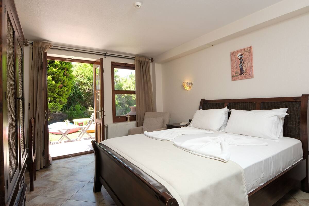 A bedroom with a large bed, a chair, a table, and a view of the outdoors through a sliding door.