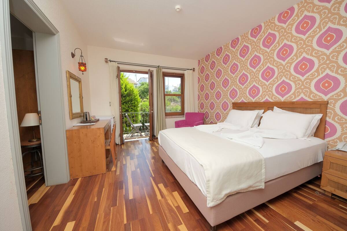 A bedroom with a wooden floor, a bed with white bedding, a wooden nightstand, a desk with a lamp, a chair, and a balcony with a view of greenery.