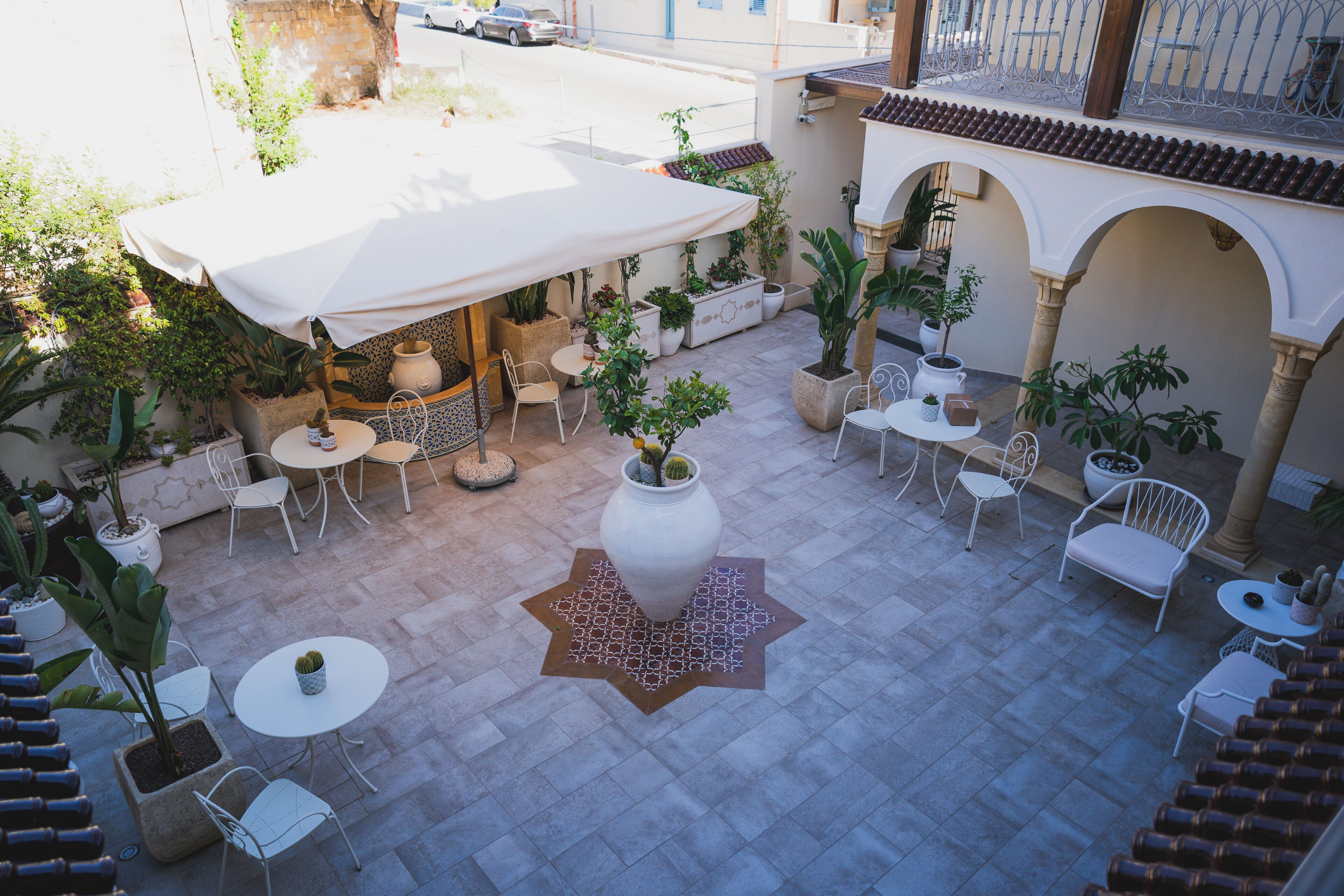 Un espace extérieur aménagé en terrasse, avec plusieurs tables et chaises, entouré de plantes en pot et doté d’un grand parasol blanc qui offre de l’ombre.