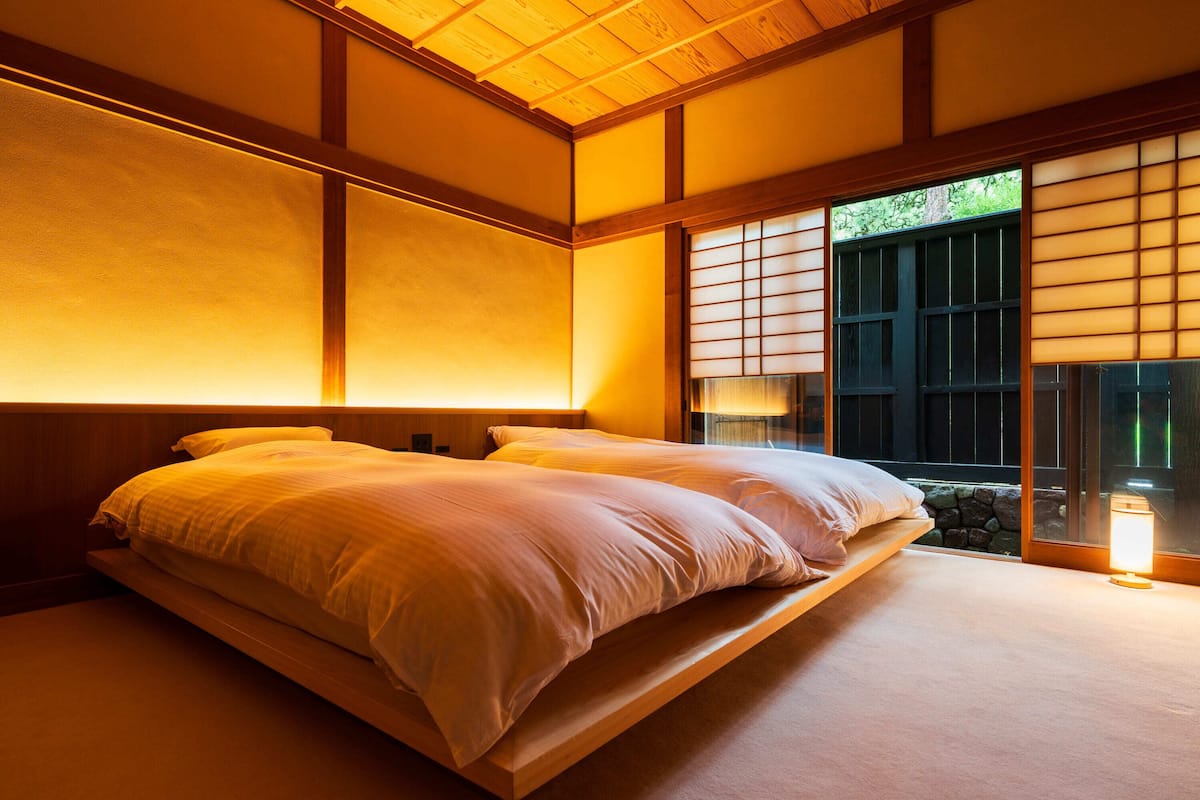 A traditional Japanese-style bedroom with tatami flooring, a low platform bed, sliding shoji doors, and a view of greenery outside.