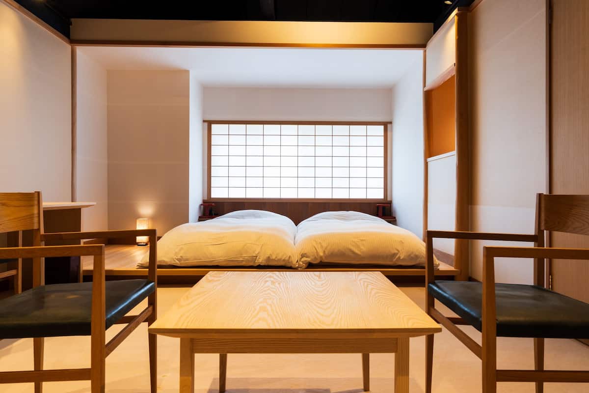 A traditional Japanese-style room with tatami flooring, a low wooden bed with a shoji screen window, and a small table with chairs.