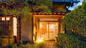 Une maison traditionnelle japonaise avec une porte en bois, un chemin de pierres et une végétation luxuriante.
