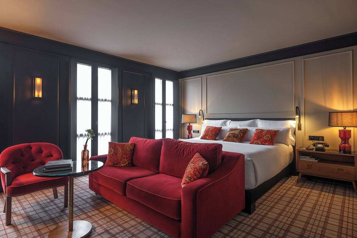 A hotel room with a red sofa, a bed with white bedding, a small table with a book, and a nightstand with a lamp.