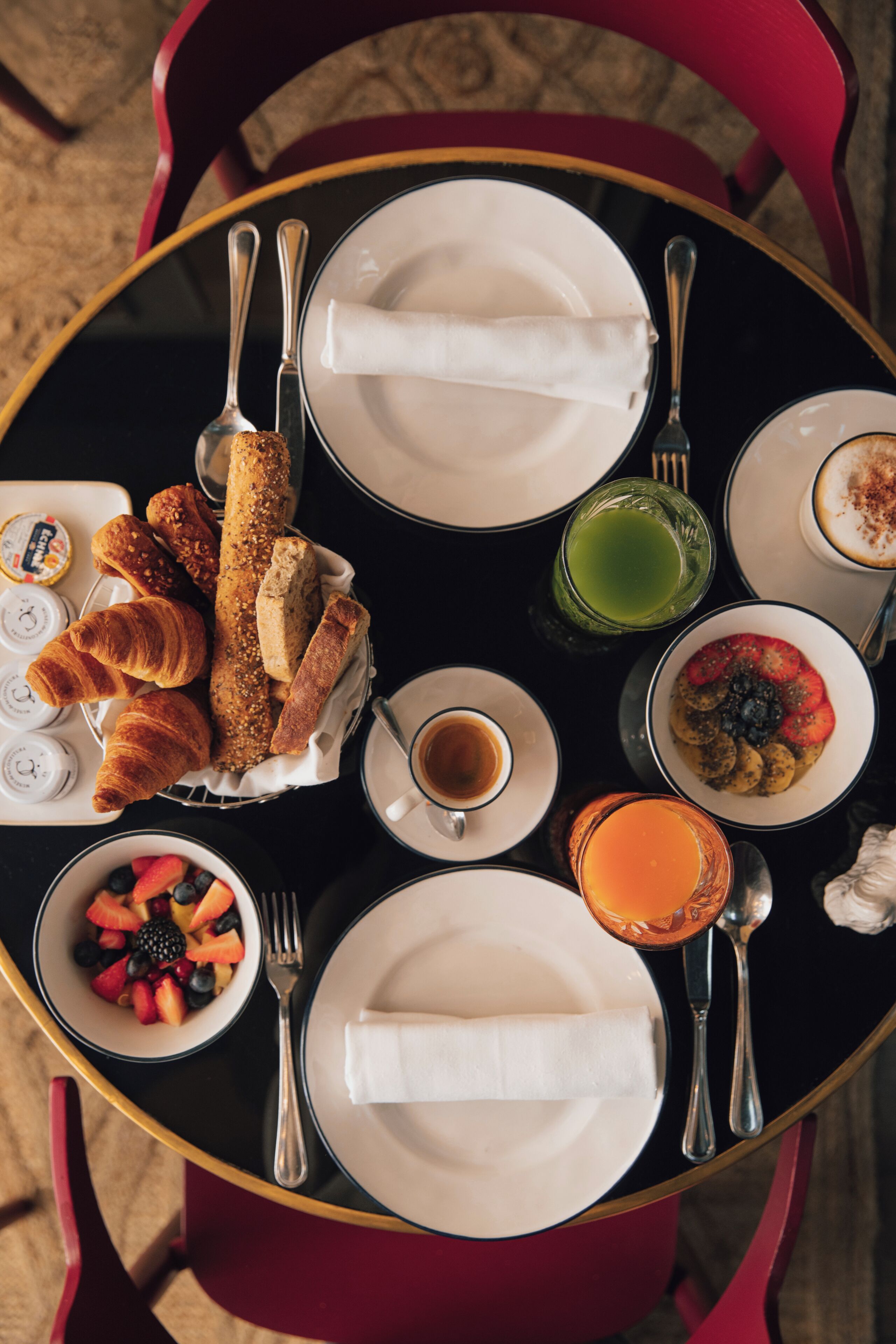 Una mesa redonda con platos, cubiertos y una variedad de alimentos, incluyendo croissants, fruta y una bebida.