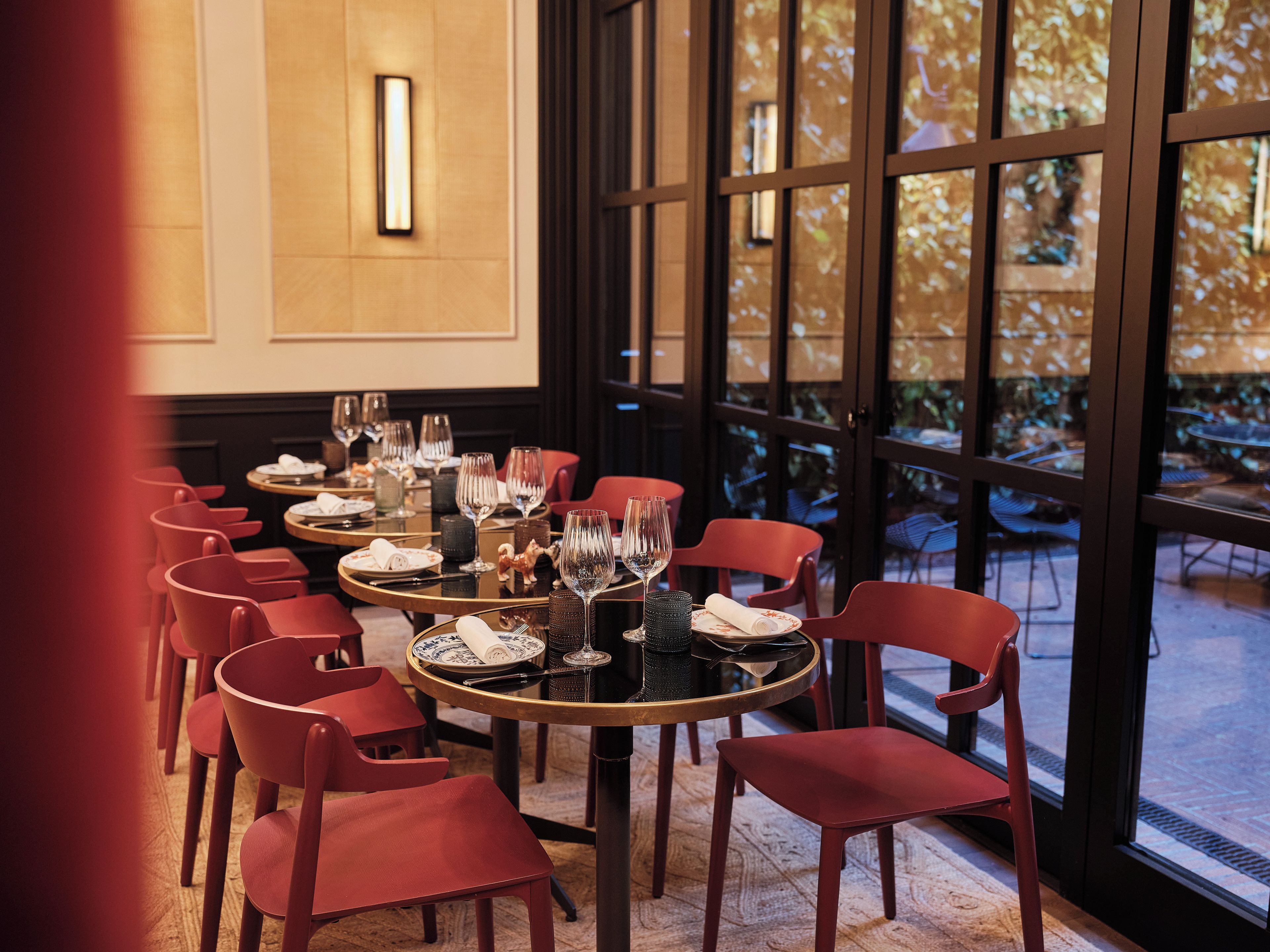 A dining area with round tables, red chairs, and a view of the outdoors through large windows.