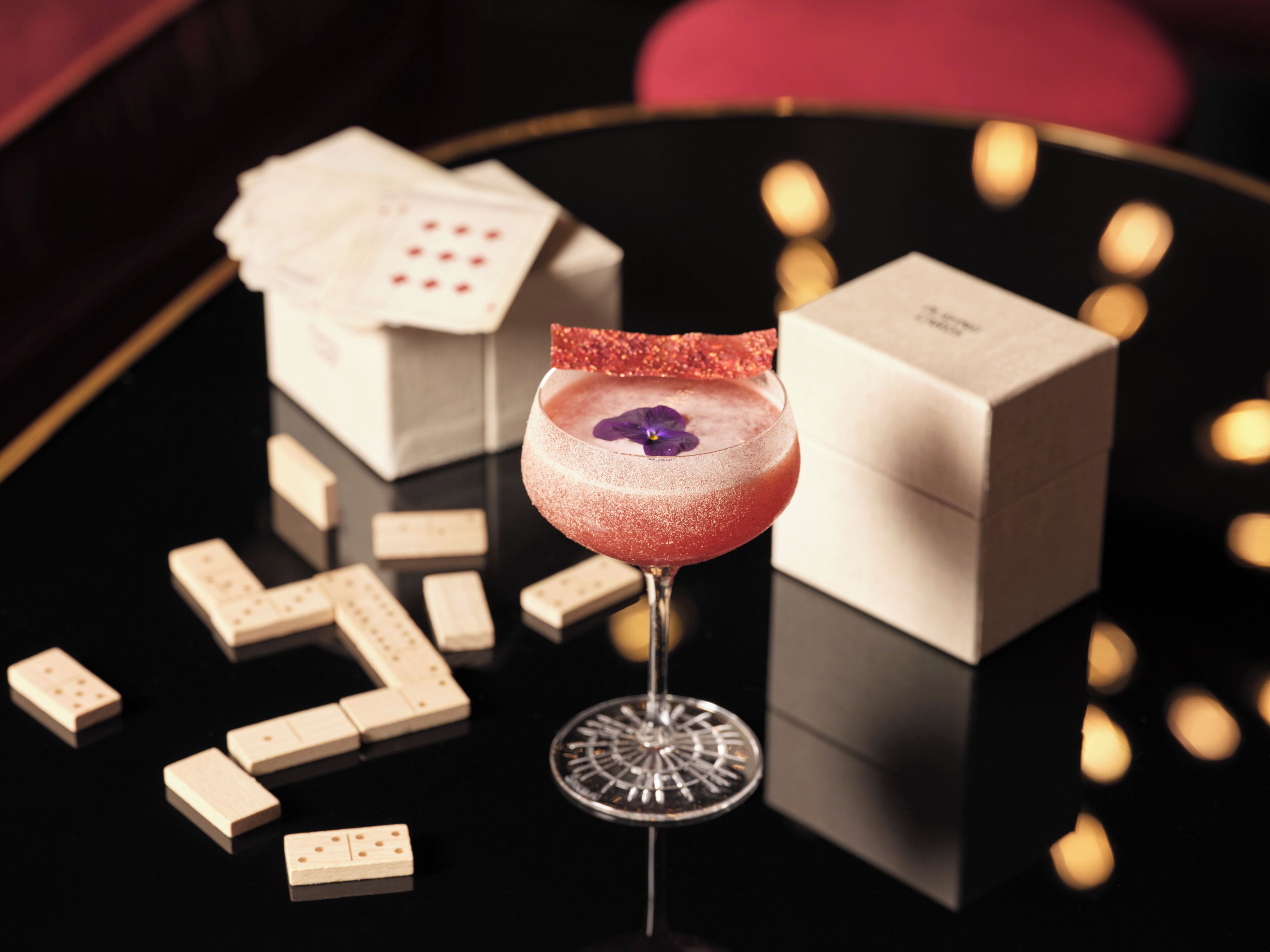 A cocktail glass with a flower garnish on a reflective surface, surrounded by dominoes and a box.