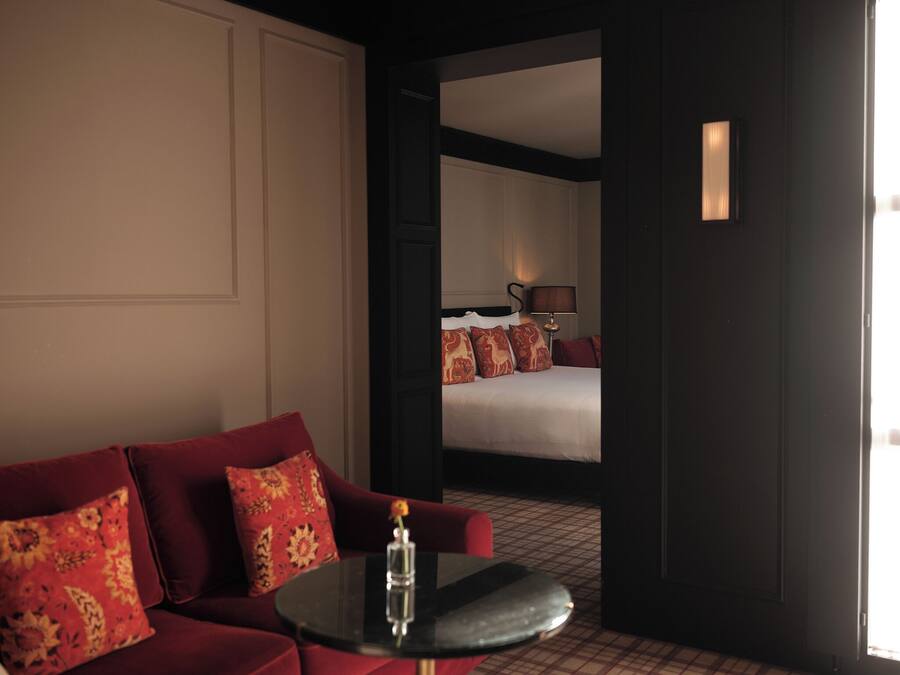 A hotel room with a red sofa, a small round table with a vase, a bed with patterned pillows, and a nightstand with a lamp.