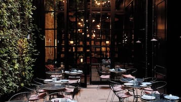 A cozy outdoor cafe with tables and chairs, surrounded by a lush green wall and a view of a warmly lit interior space.