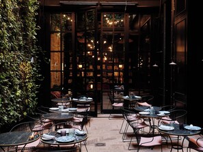 Un café extérieur accueillant, avec des tables et des chaises, entouré d’un mur verdoyant et offrant une vue sur un espace intérieur chaleureux et bien éclairé.