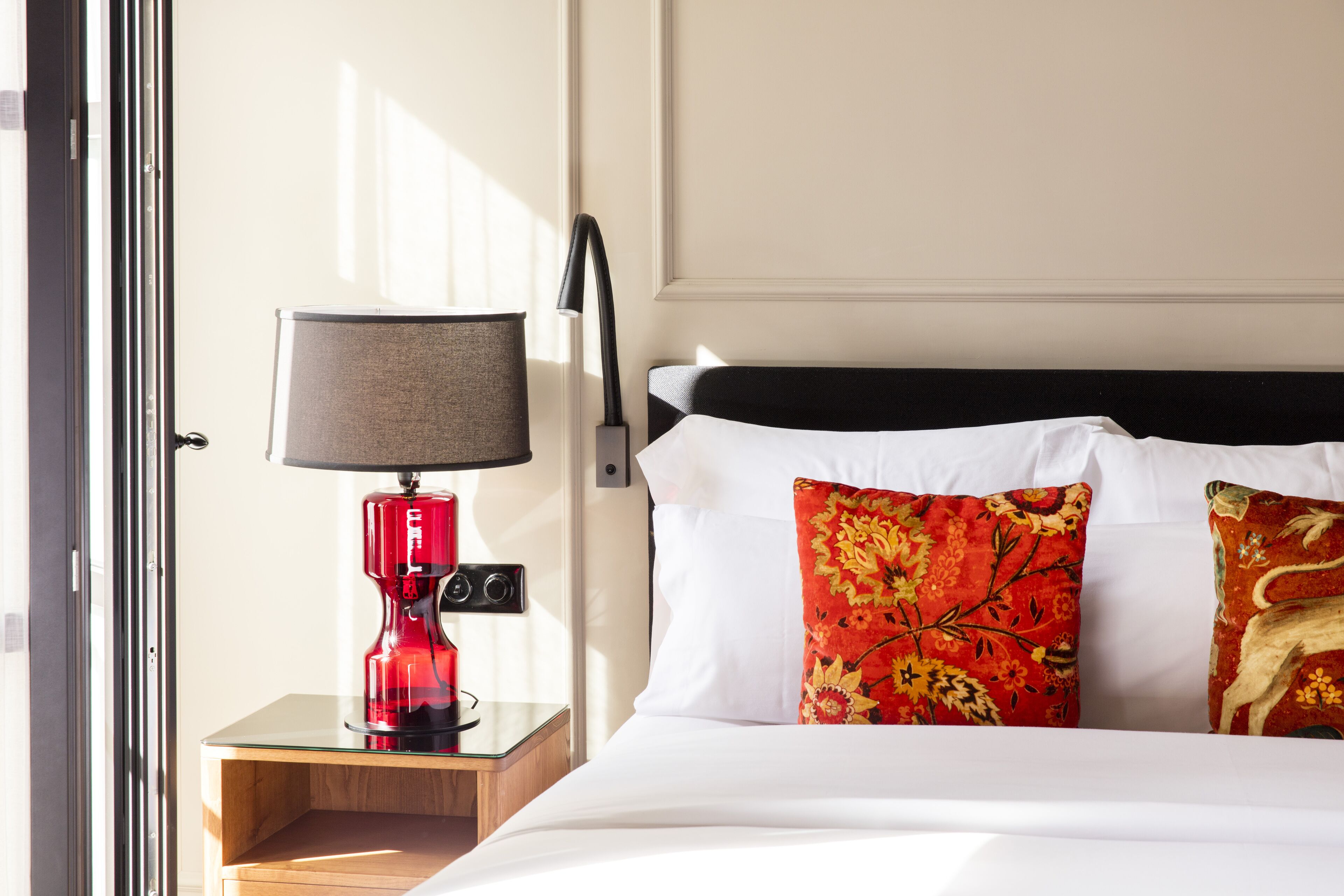A hotel room with a bed, a nightstand, a red lamp, and a window.