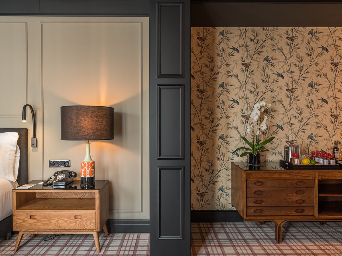 A hotel room with a bed, a nightstand with a lamp, a wooden dresser, and a floral wallpaper.