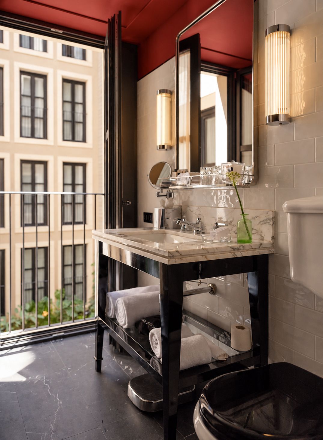 Une salle de bain moderne avec un lavabo en marbre, un grand miroir et des toilettes. Un balcon offre une vue sur les bâtiments.