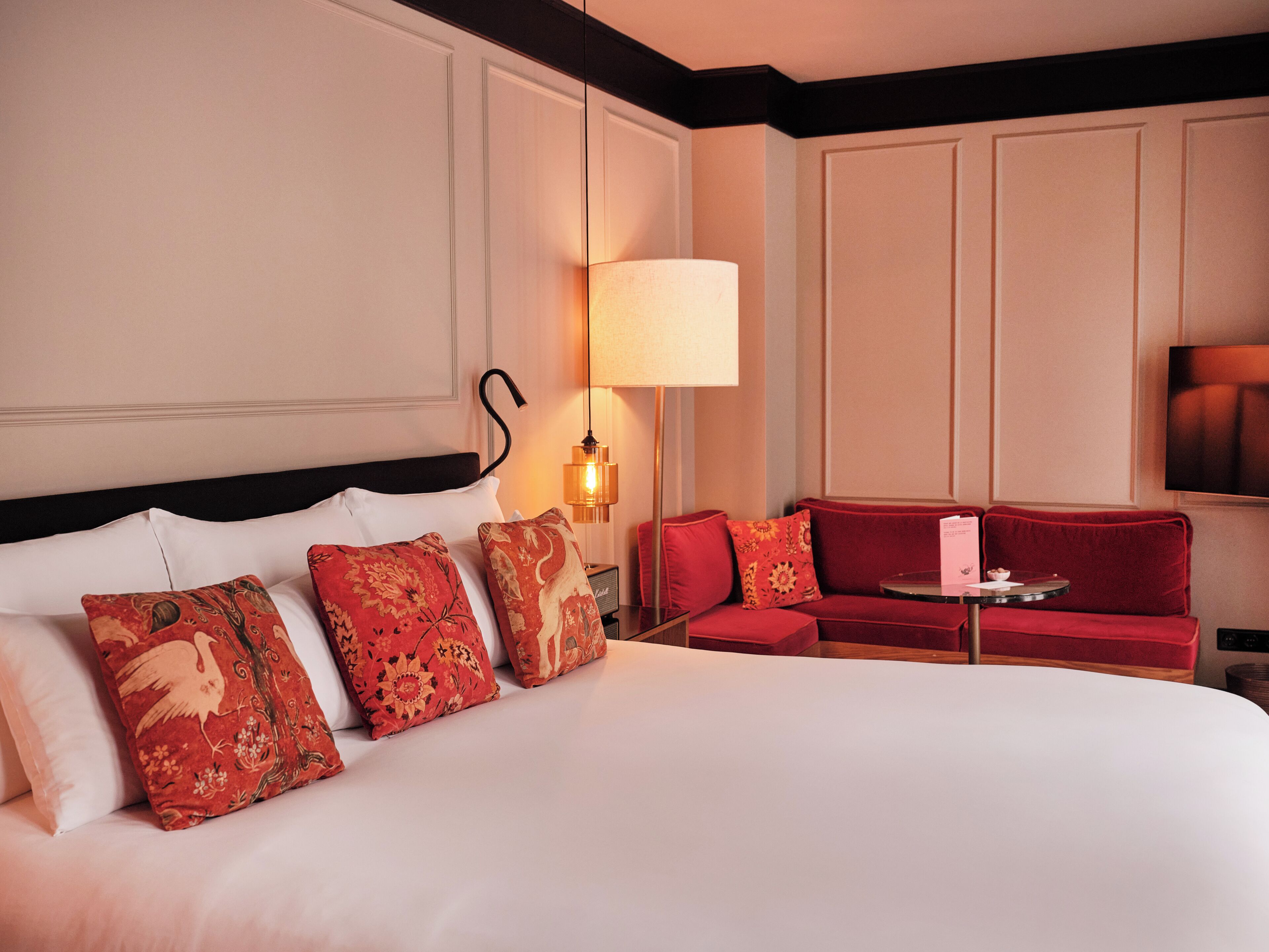 A hotel room with a large bed, a red sofa, a small table, and a wall-mounted TV.