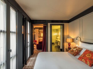 Ein Hotelzimmer mit einem Bett, einem Nachttisch mit Lampe, einem roten Sessel und Blick ins Badezimmer sowie den Flur.