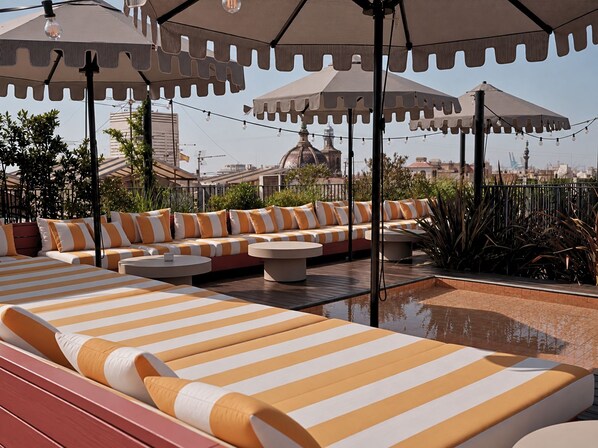 A rooftop lounge area with striped lounge chairs and ottomans, surrounded by umbrellas and string lights.