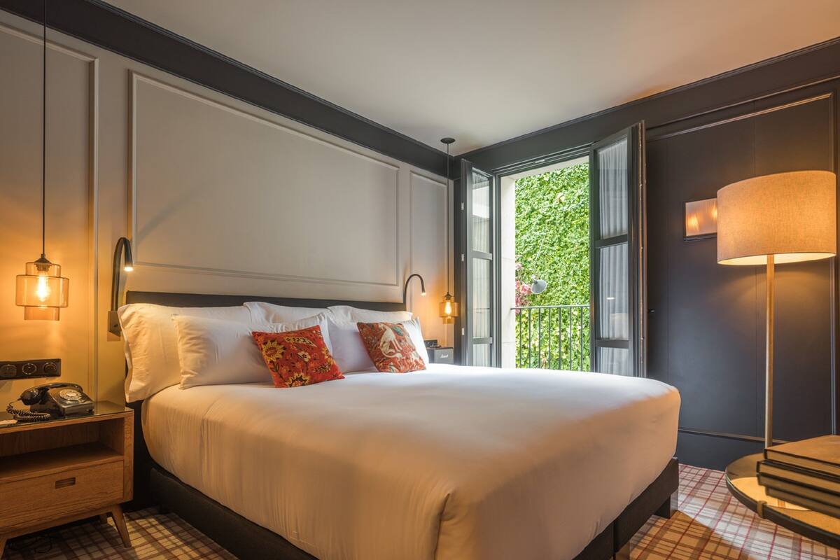 A hotel room with a large bed, a nightstand with a telephone, a lamp, and a view of greenery outside.